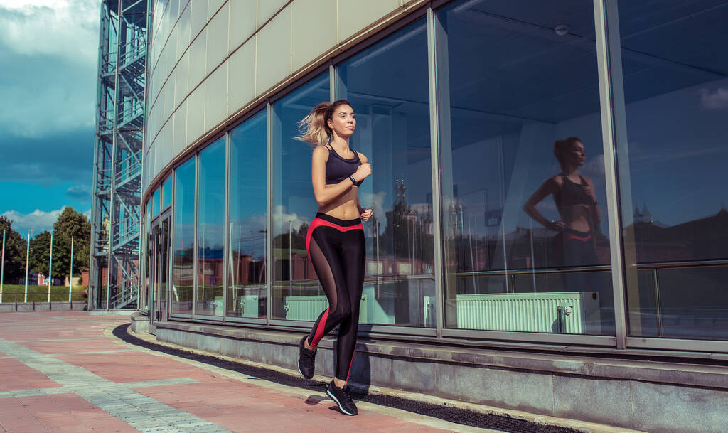 Beautiful girl, summer in city, fitness training on street, jogging in morning, sportswear, leggings top. Tanned figure. Movement in jump. Background glass windows of building. Free space copy text. - Photo, Image