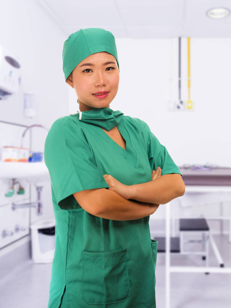 joven hermosa y feliz médico de medicina asiática o enfermera jefe del hospital en uniformes verdes y sombrero de cirujano sonriendo confiado en el fondo de la sala de la clínica
 - Foto, imagen