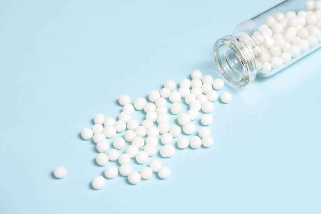 Homöopathische Globuli aus einer klaren Glasflasche auf hellblauem Hintergrund, Konzept der alternativen Homöopathie-Medizin - Foto, Bild