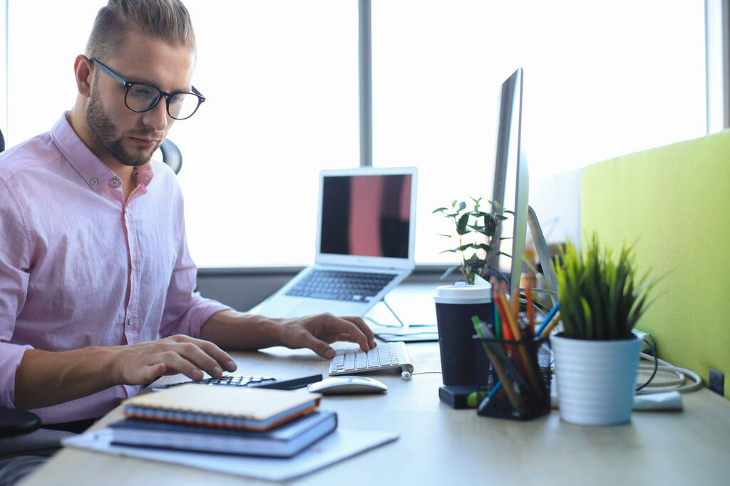 Προσεκτικός νεαρός επιχειρηματίας με πουκάμισο που εργάζεται χρησιμοποιώντας υπολογιστή ενώ κάθεται στο γραφείο. - Φωτογραφία, εικόνα