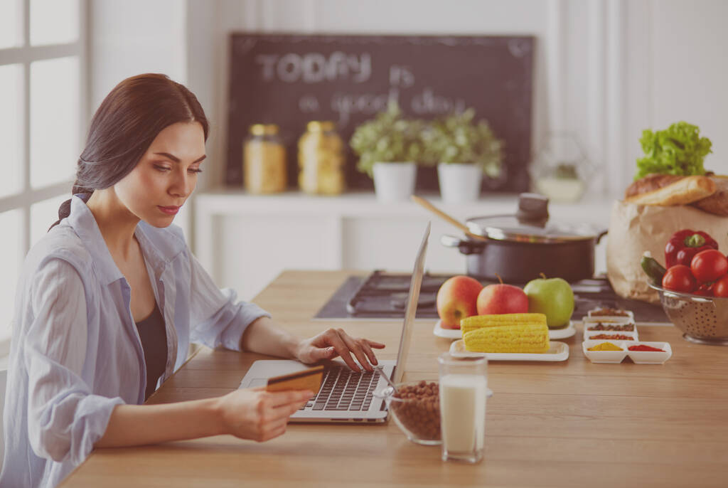 Улыбающаяся женщина совершала интернет-покупки с помощью компьютера и кредитной карты на кухне - Фото, изображение