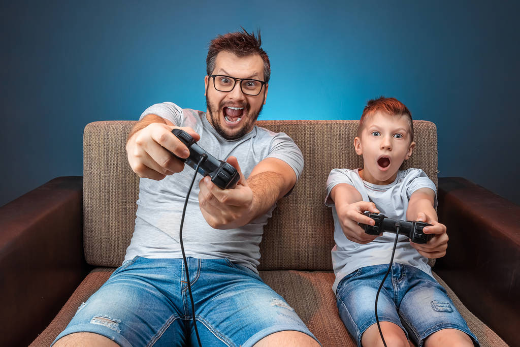 A cheerful family, dad and son play on the console, in video games, emotionally react while sitting on the couch. Day off, entertainment, leisure, spend time together. - 写真・画像