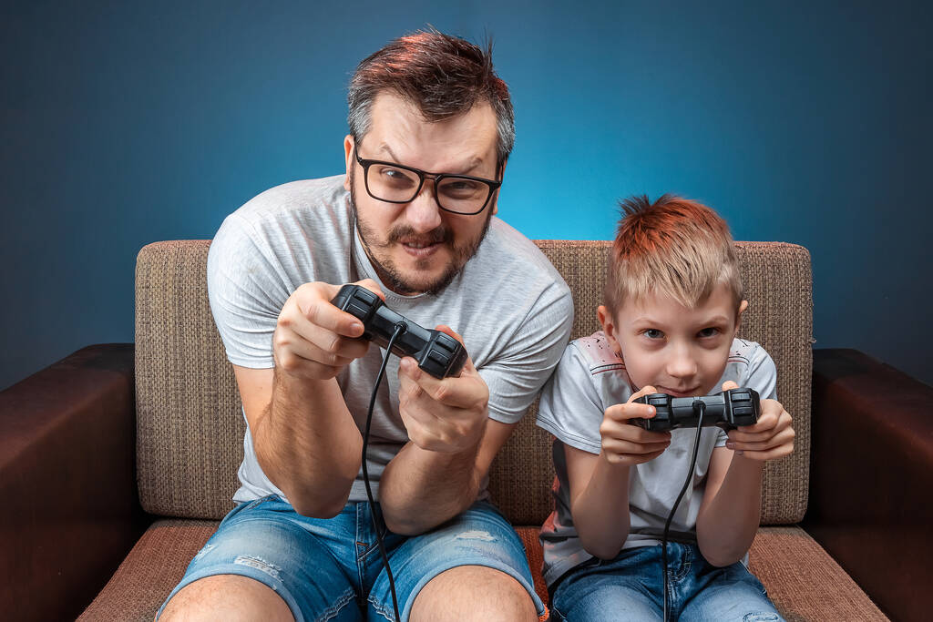 Eine fröhliche Familie, Vater und Sohn spielen an der Konsole, in Videospielen, reagieren emotional, während sie auf der Couch sitzen. Ruhetag, Unterhaltung, Freizeit, gemeinsame Zeit. - Foto, Bild