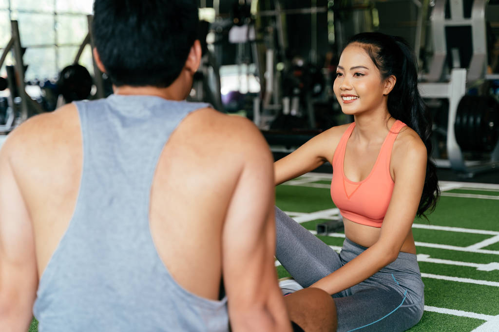 Arc fiatal ázsiai vegyes sportos nő chat egy izmos férfi ül a padlón fitnesz terem. - Fotó, kép