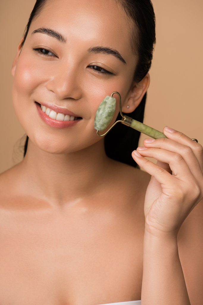 smiling beautiful naked asian girl massaging face with jade roller isolated on beige - Photo, Image