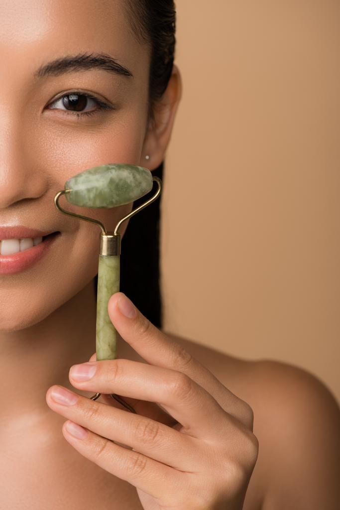 corte vista de sorrir bonito nu asiático menina massagem rosto com jade rolo isolado no bege
 - Foto, Imagem
