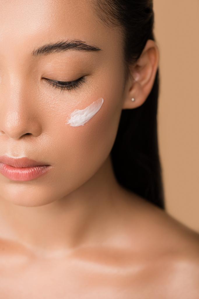 abgeschnittene Ansicht der schönen nackten asiatischen Mädchen mit Gesichtscreme auf der Wange isoliert auf beige - Foto, Bild