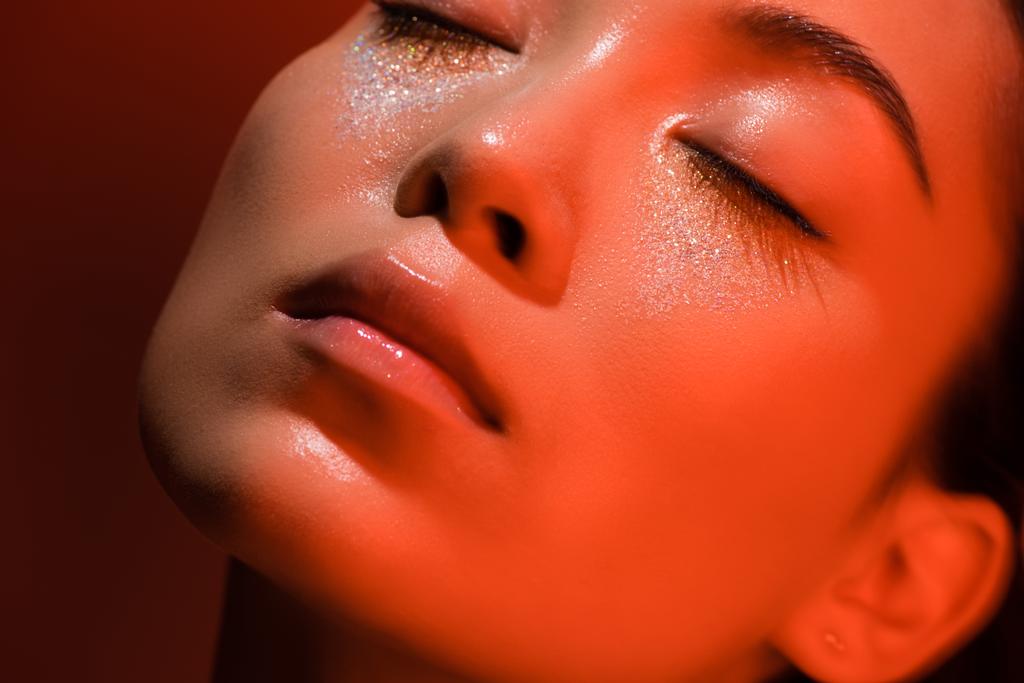 close-up uitzicht op mooie aziatische meisje met gesloten ogen en zilver schittert op het gezicht en gesloten ogen in rood licht - Foto, afbeelding