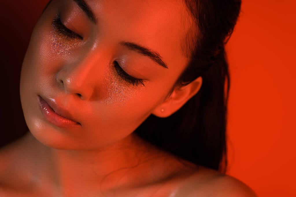 kaunis alaston aasialainen tyttö hopea kimaltelee kasvot ja suljetut silmät punaisessa valaistuksessa
 - Valokuva, kuva