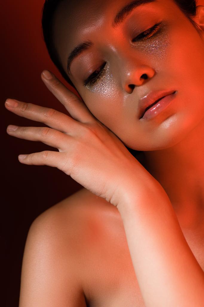 piękny nagi Azji dziewczyna z ręka w pobliżu twarz ze srebra musujące i zamknięte oczy w czerwonym oświetleniu - Zdjęcie, obraz