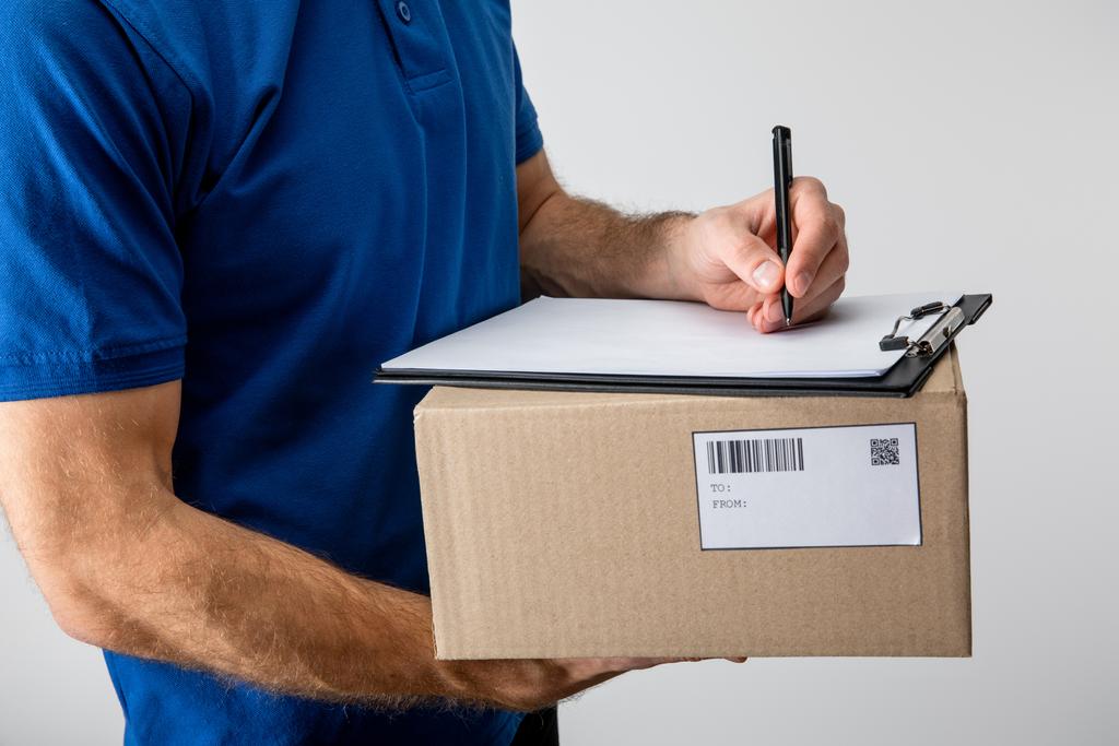 Cropped view of courier writing on clipboard and holding cardboard box isolated on grey - Photo, Image
