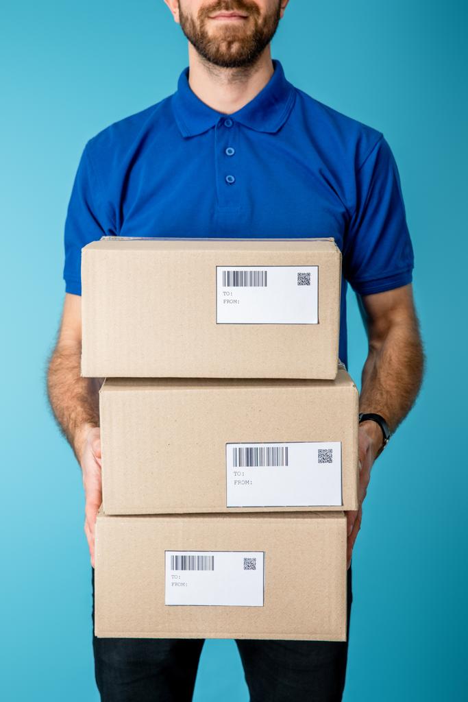 Cropped view of courier holding cardboard boxes with qr codes and barcodes on cards isolated on blue - Photo, Image