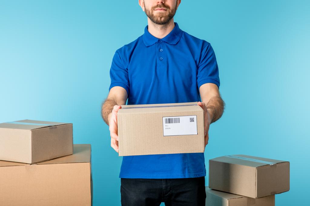 Vista recortada del mensajero que sostiene el paquete de cartón cerca de cajas aisladas en azul
 - Foto, Imagen