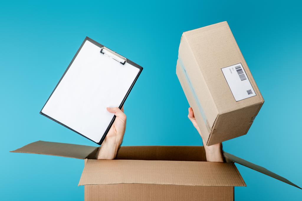 Manos de mensajero sujetando el portapapeles y el paquete de la caja de cartón aislado en azul
 - Foto, imagen