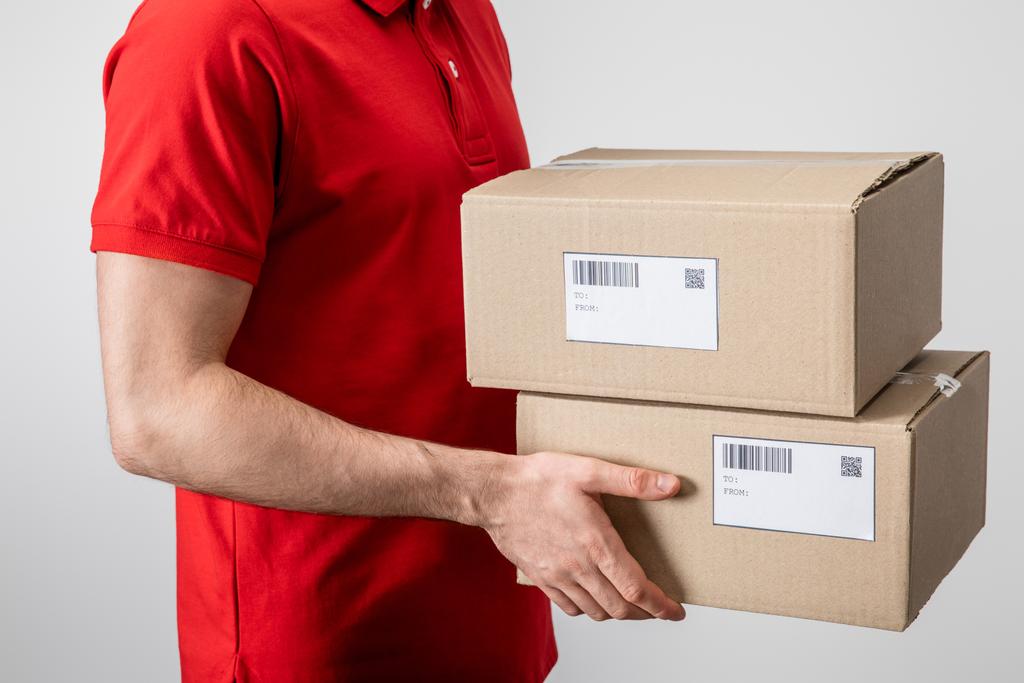 Cropped view of courier carrying cardboard packages with qr codes and barcodes on cards isolated on grey - Photo, Image