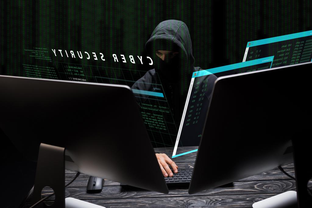hacker v masce sedí v blízkosti počítačových monitorů a kybernetické bezpečnostní písmo na černé  - Fotografie, Obrázek