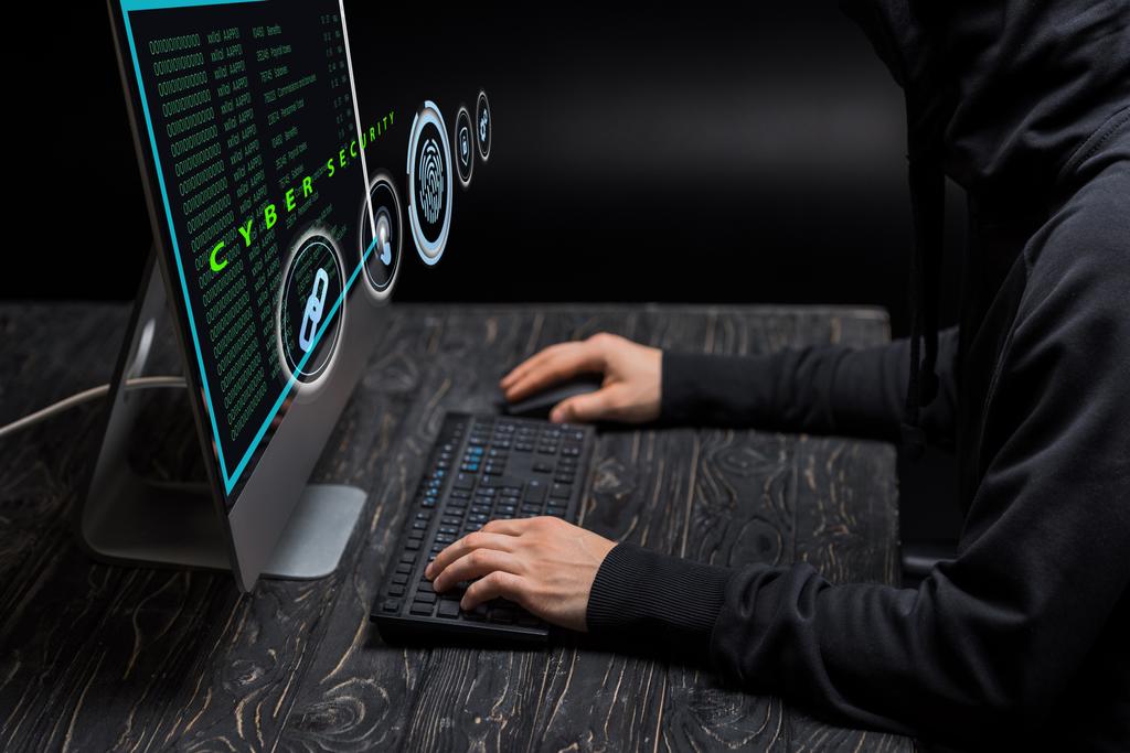 καλλιεργημένη άποψη της δακτυλογράφησης χάκερ στο πληκτρολόγιο του υπολογιστή κοντά στην οθόνη του υπολογιστή με ηλεκτρονικά γράμματα ασφαλείας στο μαύρο  - Φωτογραφία, εικόνα