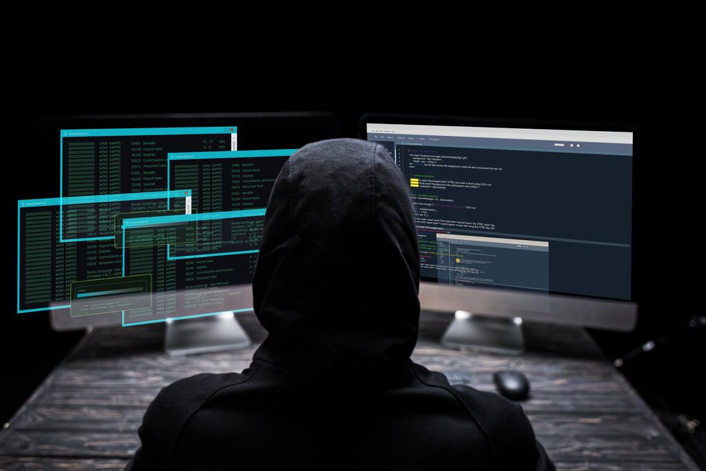 πίσω όψη κουκουλοφόρου χάκερ που κάθεται κοντά σε οθόνες υπολογιστών με δεδομένα σε οθόνες σε μαύρο  - Φωτογραφία, εικόνα