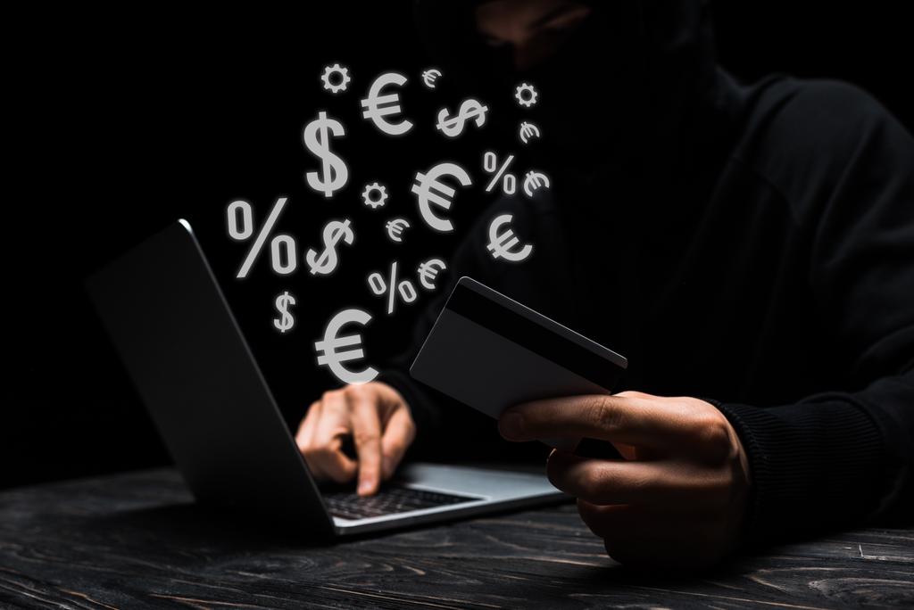 selective focus of hacker in mask using laptop while holding credit card near money signs on black  - Photo, Image