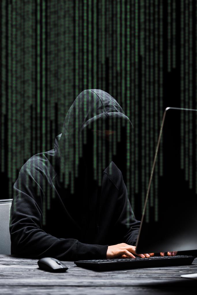 hacker in mask sitting near computer monitor and typing on computer keyboard  near illustration on black  - Photo, Image