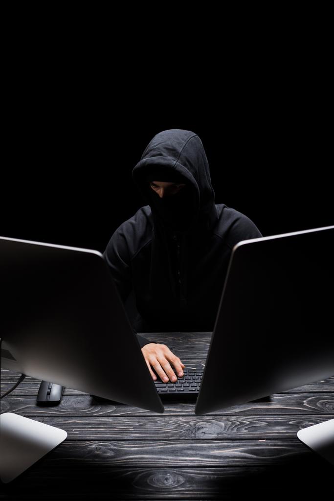hombre encapuchado en la máscara sentado cerca de monitores de ordenador aislado en negro
  - Foto, Imagen