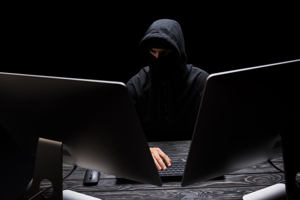 χάκερ με μάσκα που κάθεται κοντά σε οθόνες υπολογιστών που απομονώνονται σε μαύρο  - Φωτογραφία, εικόνα