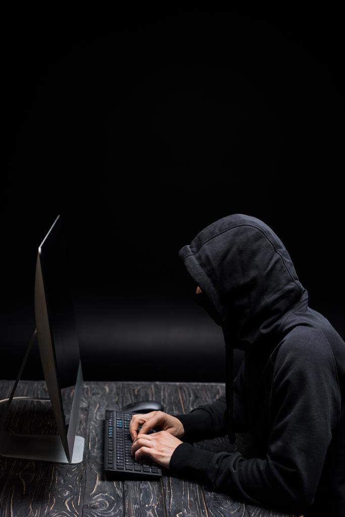 Siyah üzerine bilgisayar klavyesinde yazan maskeli bir bilgisayar korsanı.  - Fotoğraf, Görsel