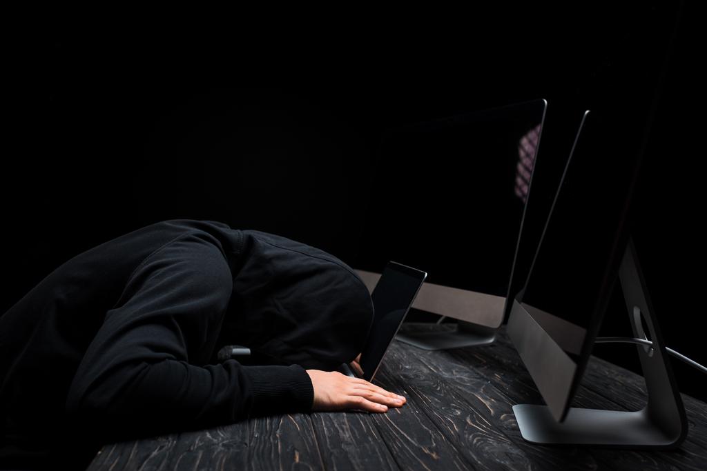 zdenerwowany haker leżący na laptopie w pobliżu monitorów komputerowych z pustym ekranem izolowanym na czarno  - Zdjęcie, obraz
