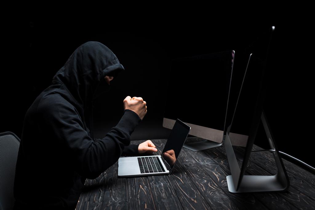 hacker enojado con puño cerrado cerca de la computadora portátil y monitores de ordenador con pantalla en blanco aislado en negro
  - Foto, Imagen