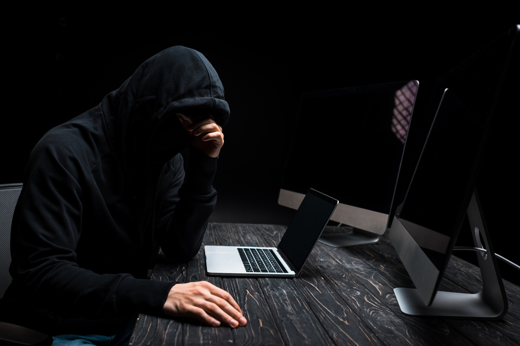 rozrušený hacker sedí v blízkosti notebooku a počítačové monitory s prázdnou obrazovkou izolované na černé  - Fotografie, Obrázek