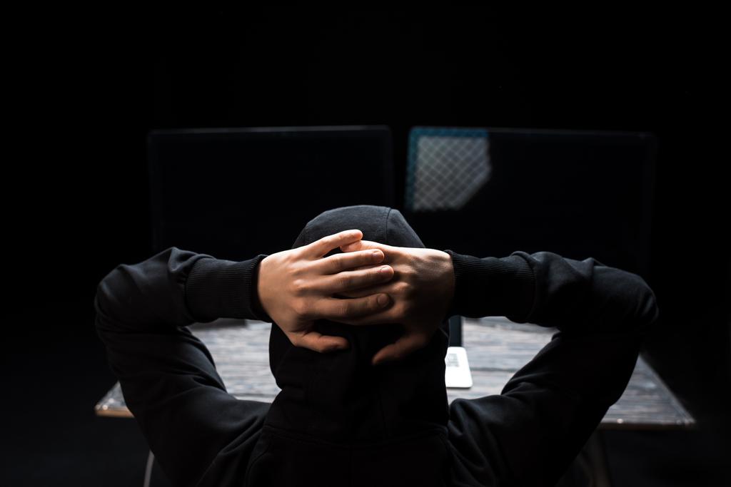 vista posterior de hacker con capucha con las manos detrás de la cabeza cerca de monitores de computadora con pantalla en blanco aislado en negro
  - Foto, Imagen