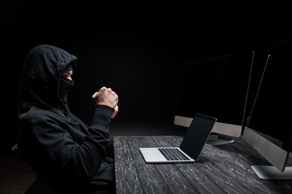 hacker met gebalde vuisten op zoek naar laptop met leeg scherm in de buurt van computer monitoren geïsoleerd op zwart  - Foto, afbeelding