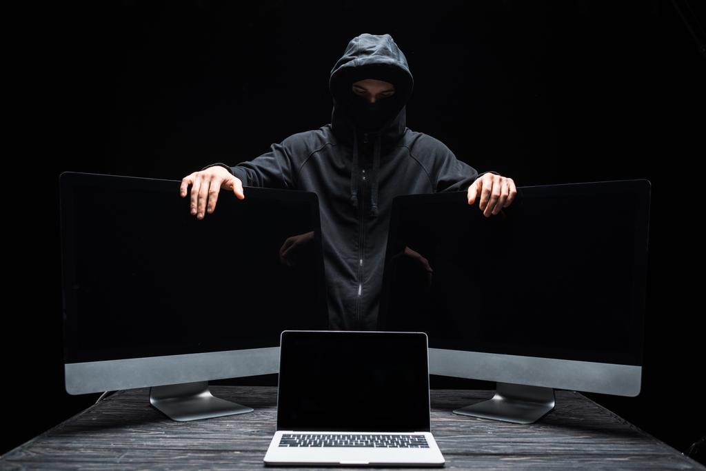 hacker in masker aanraken computer monitoren in de buurt van laptop met blanco scherm geïsoleerd op zwart  - Foto, afbeelding