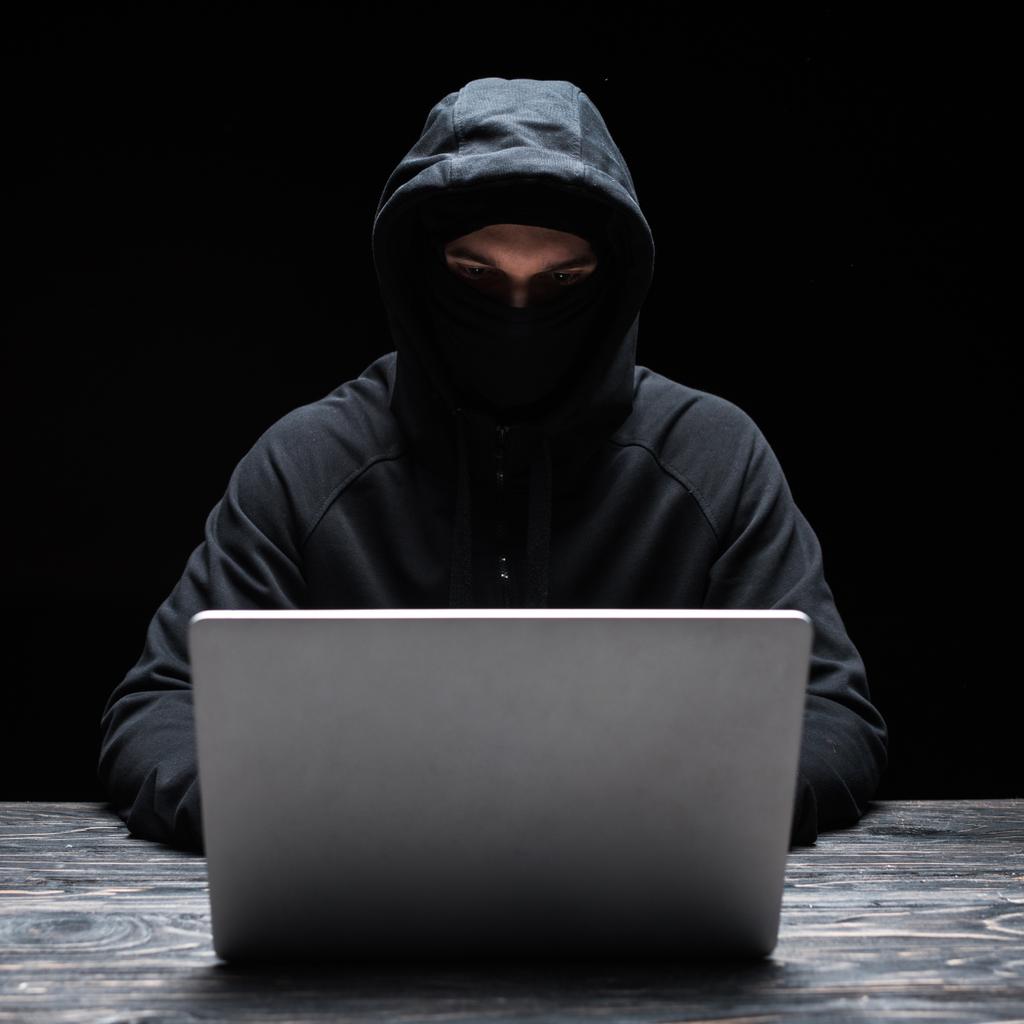 νεαρός χάκερ με μάσκα και κουκούλα χρησιμοποιώντας φορητό υπολογιστή απομονωμένο σε μαύρο  - Φωτογραφία, εικόνα