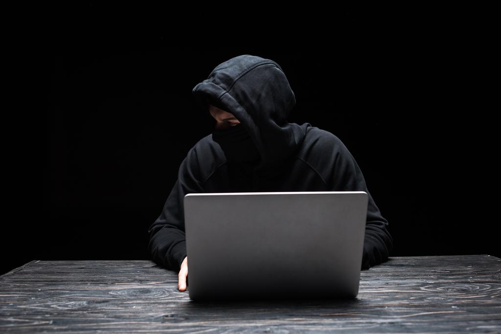 młody haker w masce patrząc daleko podczas korzystania z laptopa izolowane na czarno  - Zdjęcie, obraz