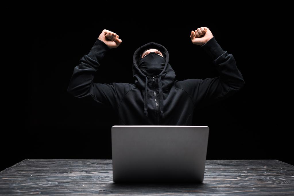 схвильований хакер в масці святкування тріумфу біля ноутбука ізольовано на чорному
  - Фото, зображення