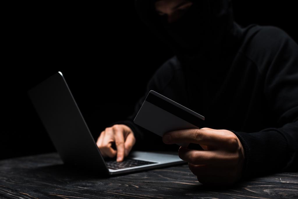 enfoque selectivo de hacker en la máscara usando el ordenador portátil mientras mantiene la tarjeta de crédito aislada en negro
  - Foto, imagen