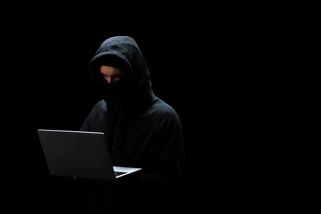 Siyah üzerine izole edilmiş dizüstü bilgisayar kullanan maskeli ve başlıklı bilgisayar korsanı  - Fotoğraf, Görsel