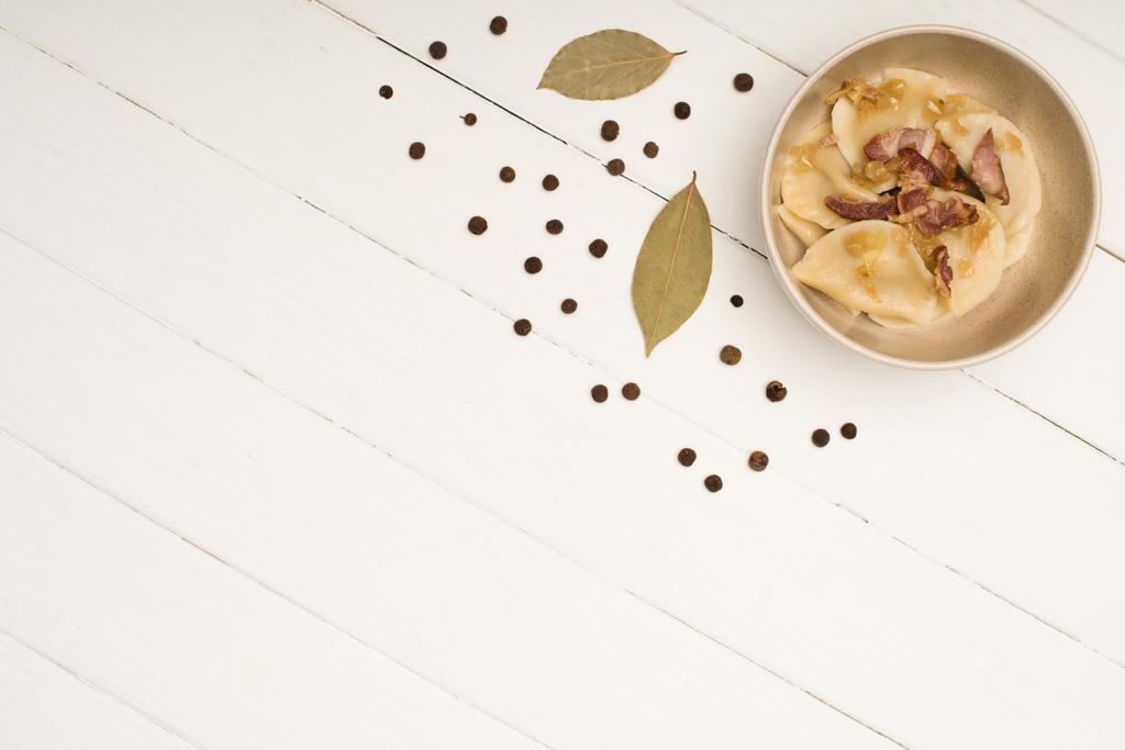 vrchní pohled na lahodné varenyky s prasklinami v misce v blízkosti bobkový listy a černý peppercorns na bílém dřevěném stole - Fotografie, Obrázek