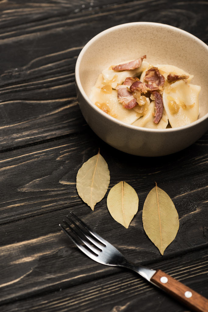 delicious varenyky with cracklings in bowl near bay leaves and fork on black wooden table - 写真・画像