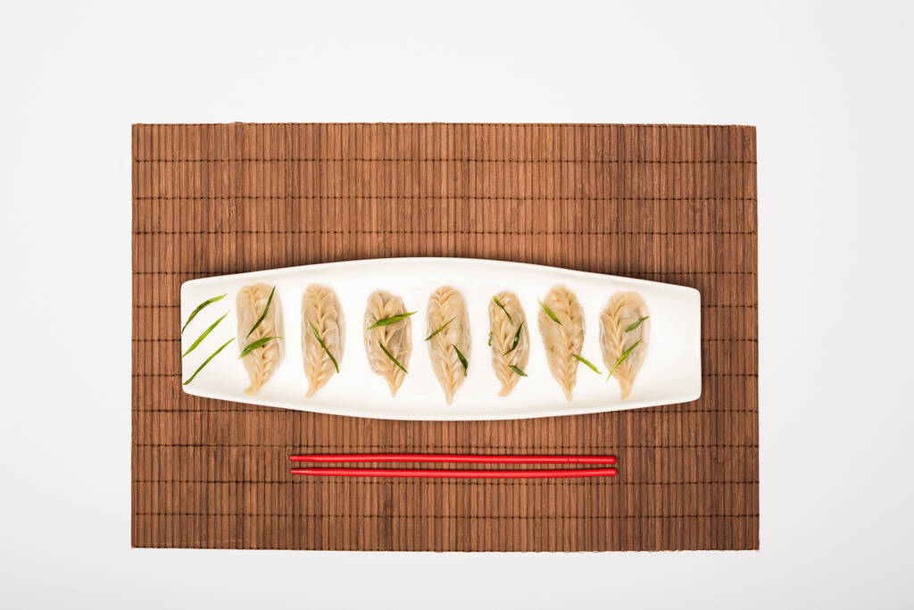 bovenaanzicht van heerlijke Chinese gekookte knoedels op bord bij eetstokjes op bamboe mat op witte achtergrond - Foto, afbeelding
