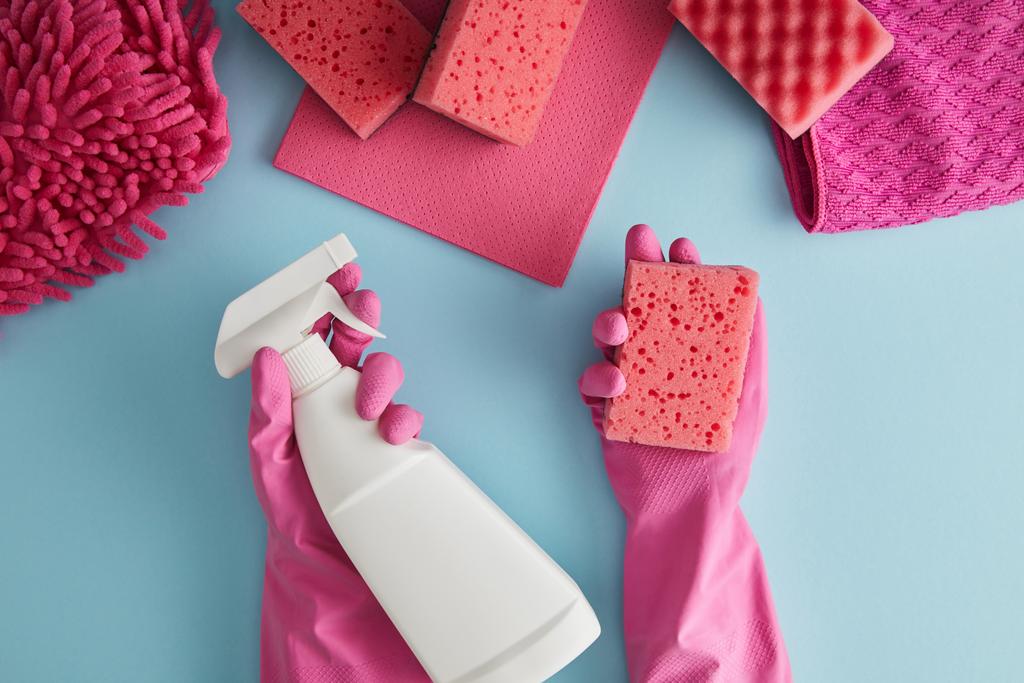 bijgesneden weergave van huishoudster in roze rubberen handschoenen met spons en spuitfles op blauw met vodden - Foto, afbeelding