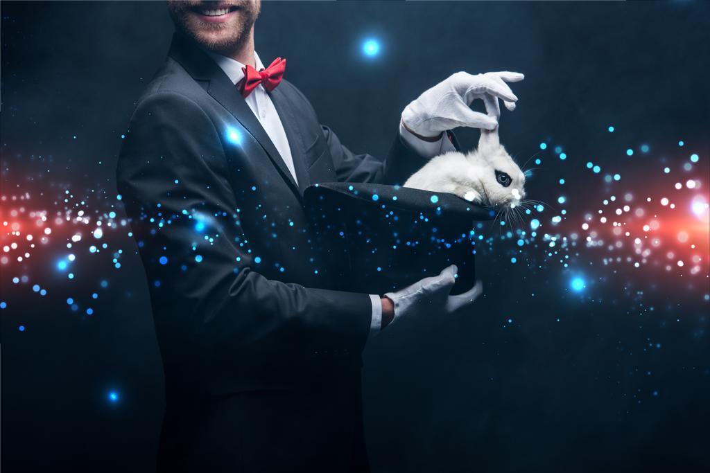abgeschnittene Ansicht eines lächelnden Zauberers, der einen Trick mit weißem Kaninchen im Hut zeigt, in einem dunklen Raum mit Rauch und glühender Illustration - Foto, Bild