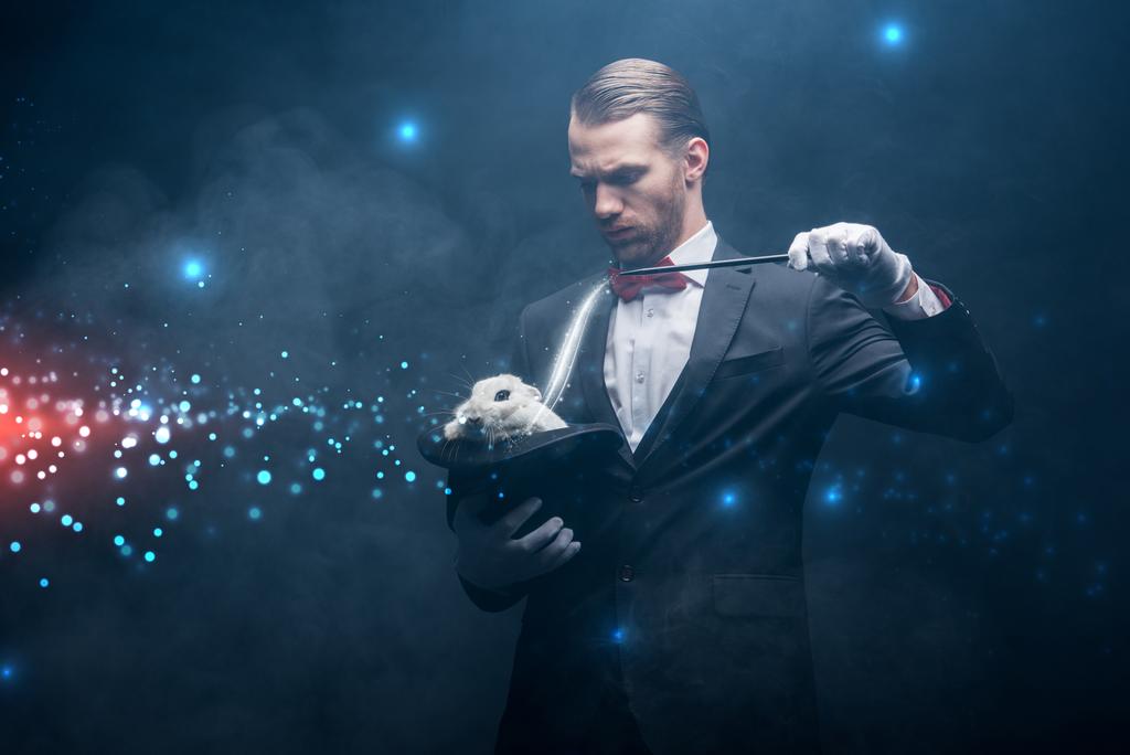 seriózní kouzelník v obleku předváděcí trik s hůlkou a bílým králíkem v klobouku, tmavý pokoj s kouřem a zářící ilustrací - Fotografie, Obrázek