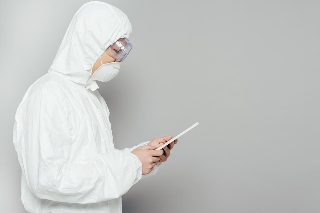 side view of asian epidemiologist in hazmat suit and respirator mask using digital tablet on grey background - Photo, Image