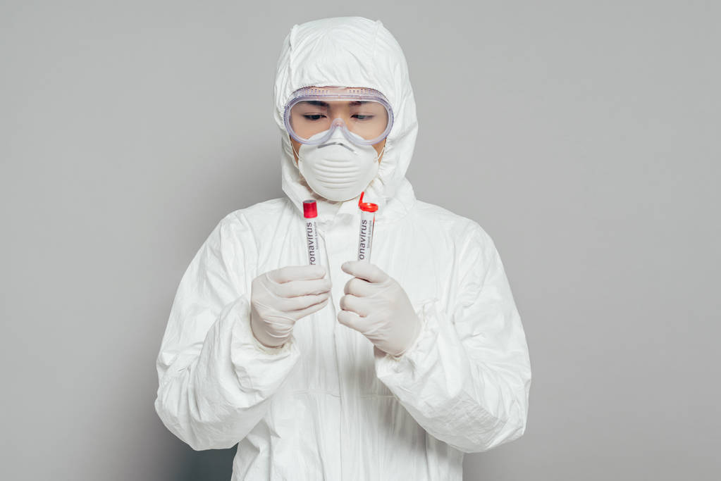 Tehlikeli madde giysisi ve solunum maskesi takmış Asyalı epidemiyoloji uzmanı gri arka planda kan örnekleri olan test tüpleri tutuyor. - Fotoğraf, Görsel