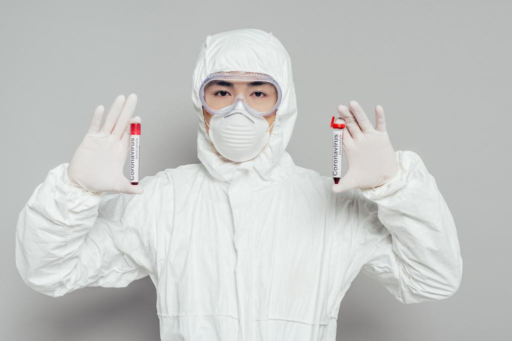 Ασιάτης επιδημιολόγος με στολή και μάσκα αναπνοής που δείχνει δοκιμαστικούς σωλήνες με δείγματα αίματος σε γκρι φόντο - Φωτογραφία, εικόνα