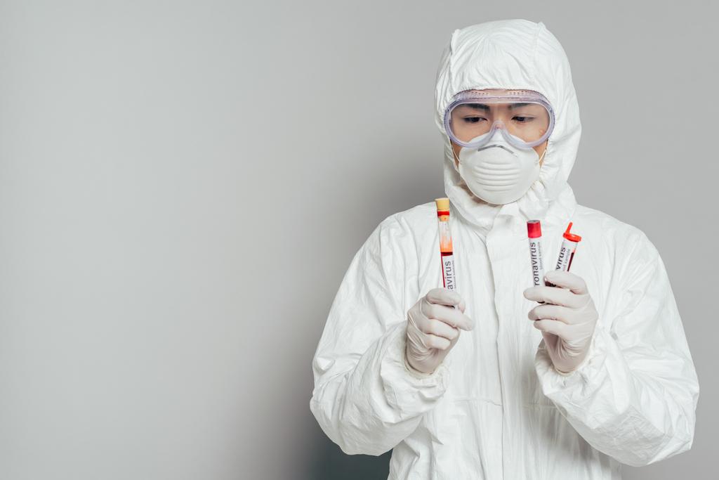 aziatische epidemioloog in hazmat pak en masker houden reageerbuizen met bloedmonsters op grijze achtergrond - Foto, afbeelding