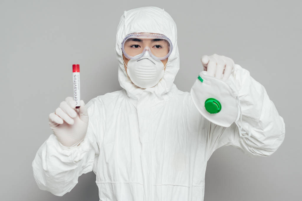 Asiatischer Epidemiologe im Warnanzug mit Atemschutzmaske und Reagenzglas mit Blutprobe auf grauem Hintergrund - Foto, Bild