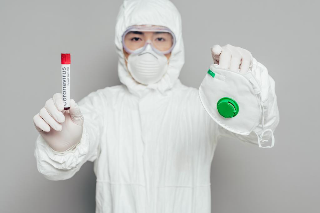 azjatycki epidemiolog w kombinezonie ochronnym patrzący w kamerę trzymający maskę respiratora i probówkę z próbką krwi na szarym tle - Zdjęcie, obraz